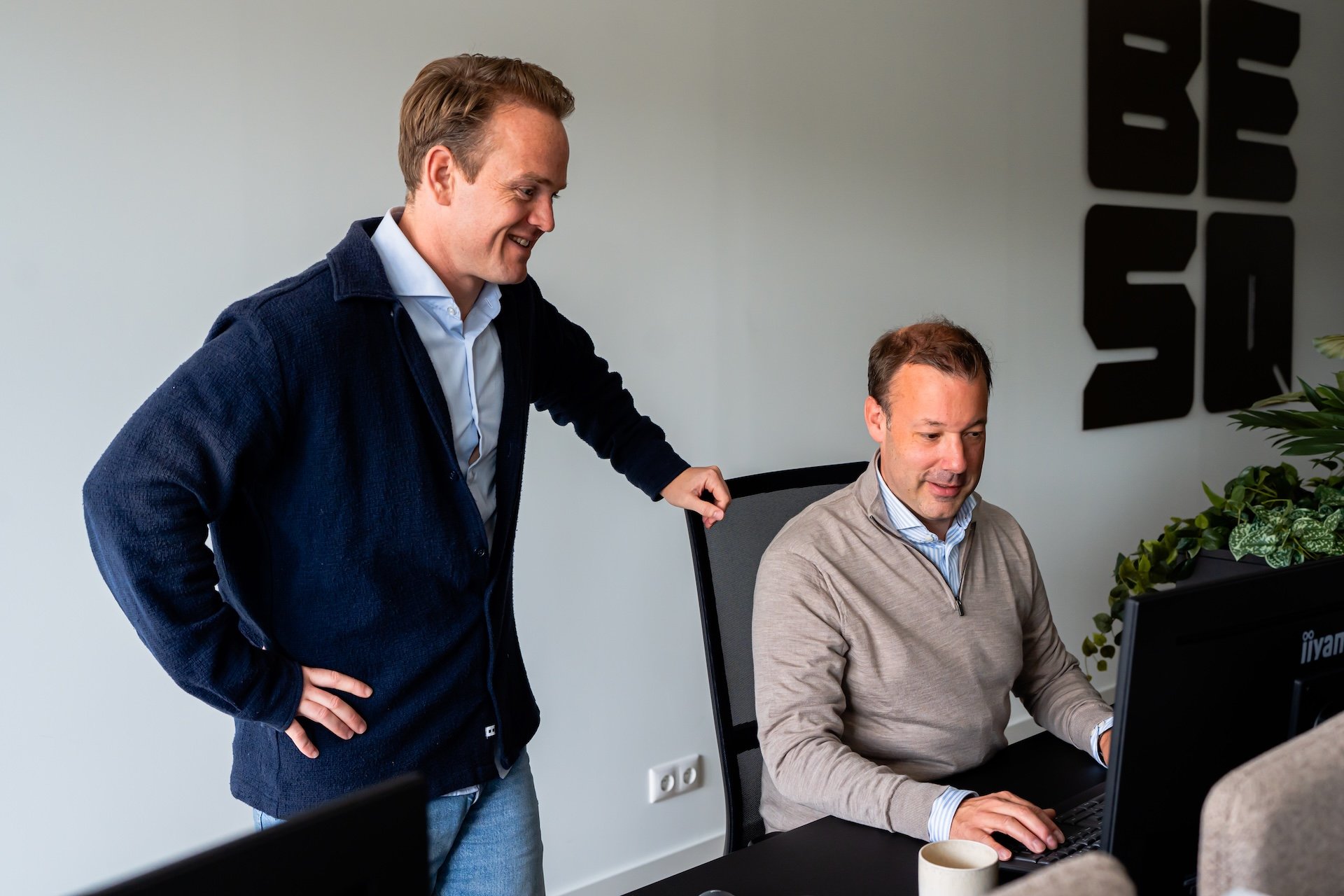 Twee mannelijke collega's overleggen lachend samen achter een computer, met het logo van BESQ Group op de muur in de achtergrond.